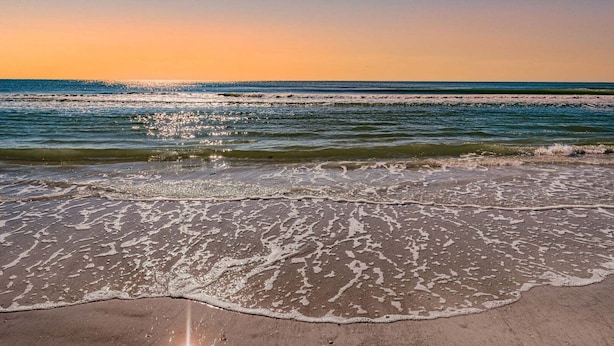 Ein Strand mit Wellen, die ans Ufer rollen, einem klaren Himmel und einem Sonnenuntergang.