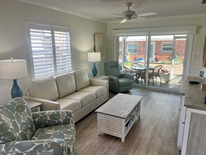A living room with a sofa, armchair, coffee table, and a lamp. There is a sliding glass door leading to an outdoor patio area.