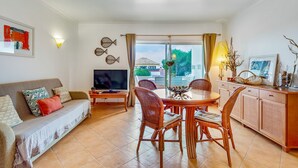 A living room with a sofa, TV, dining table, and chairs. There is a window with curtains, a wall-mounted TV, and a view of a building outside. - Golden 1 BDR Flat W/ Balcony & Pool by LovelyStay (Cabanas)