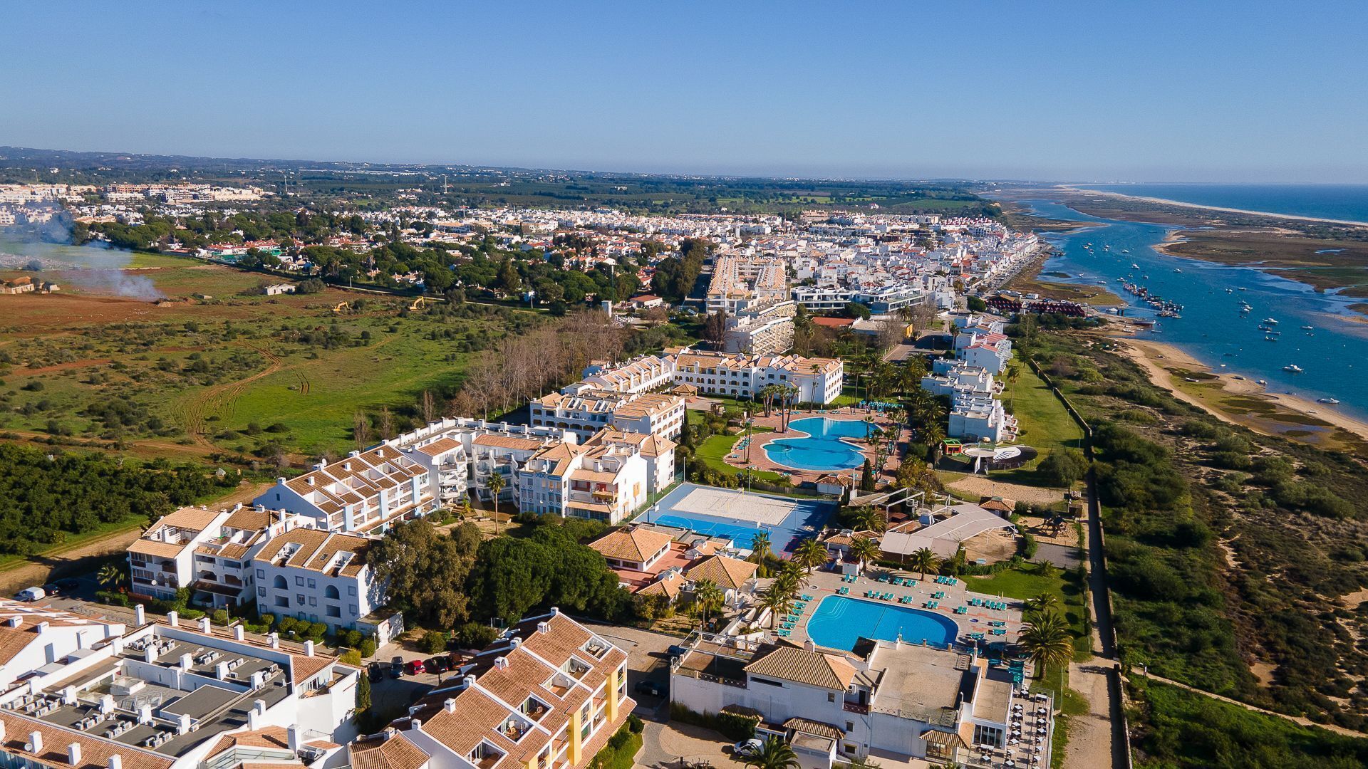 An aerial view of a coastal resort with multiple buildings, a large swimming pool, and a beach area.