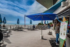 A wooden deck with picnic tables and blue umbrellas, overlooking a beach.