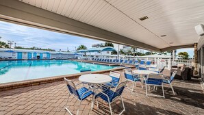 A covered swimming pool area with lounge chairs and tables, a clear view of the pool, and a building with a blue roof in the background.