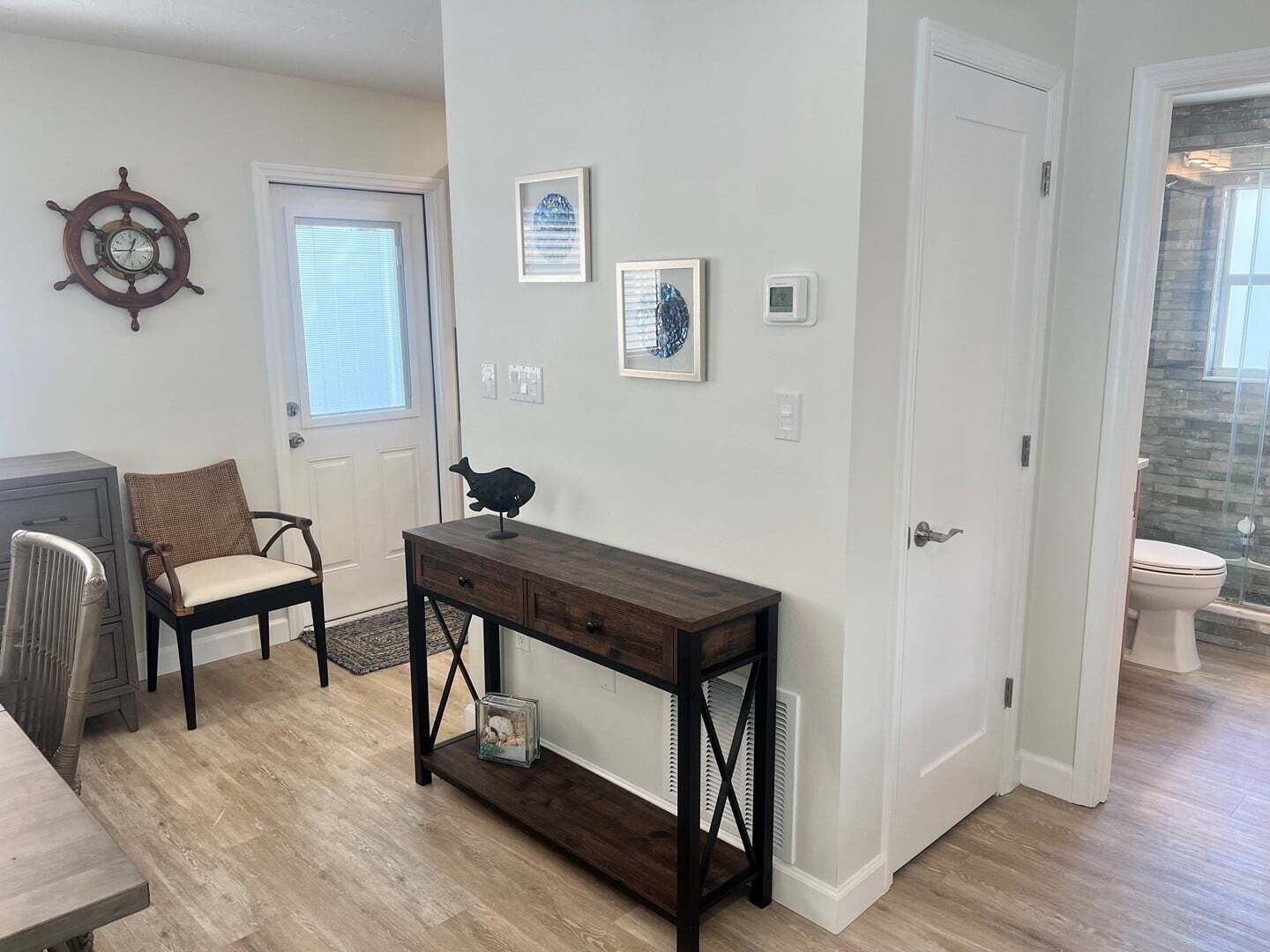 Una habitación con piso de madera, una consola de madera oscura, una silla, un objeto decorativo pequeño sobre la mesa, un timón de barco montado en la pared, y una puerta que da a otra habitación.