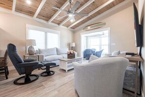 Ein geräumiges Wohnzimmer mit hoher Decke, Holzbalken, Deckenventilator und einem großen Fenster, das für natürliches Licht sorgt.