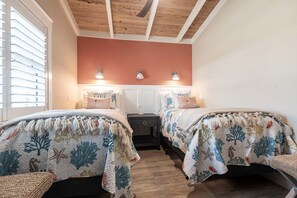 A bedroom with two beds, each with a beach-themed bedspread, a wooden ceiling, and a window with blinds.