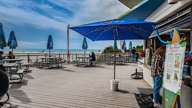 Una terraza de madera con mesas de picnic y sombrillas azules, con vistas a una playa.