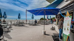 A wooden deck with picnic tables and blue umbrellas, overlooking a beach.