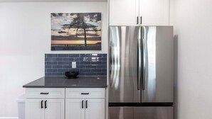 A kitchen with a stainless steel refrigerator, white cabinets, a black countertop, and a sunset painting on the wall.
