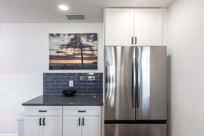 Une cuisine dotée d’un réfrigérateur en acier inoxydable, de meubles blancs, d’un plan de travail noir et d’un tableau représentant un coucher de soleil accroché au mur.
