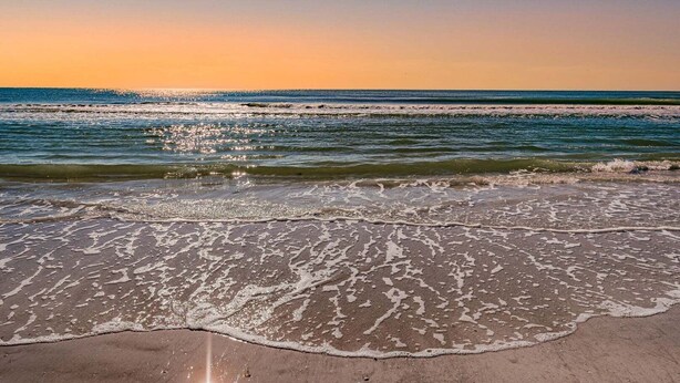 Een strand met golven die de kust bereiken, een heldere lucht en een ondergaande zon.