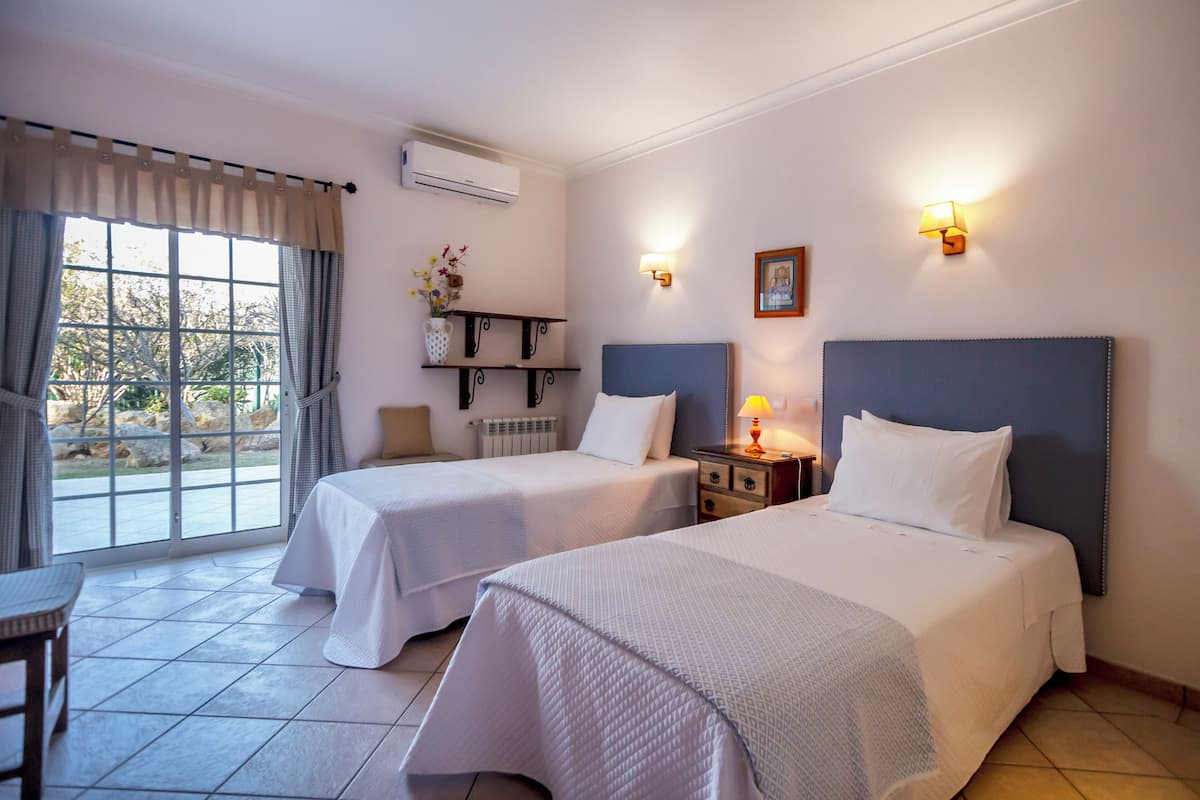 A hotel room with two beds, a dining table, a window with curtains, a wall-mounted air conditioner, and a small shelf with decorative items.