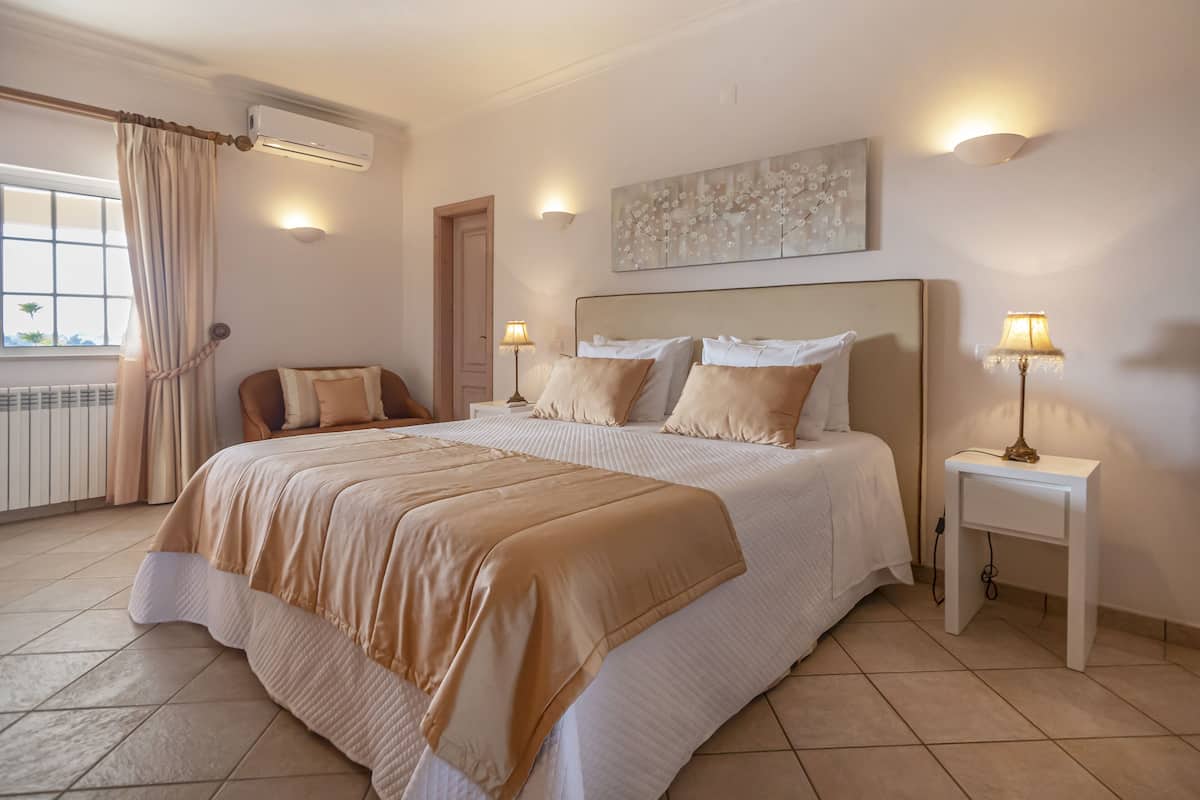 A bedroom with a large bed, two bedside tables, a window with curtains, and a wall-mounted air conditioner.