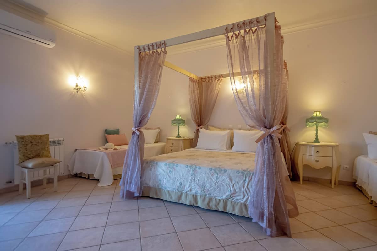 A four-poster bed with sheer curtains, a bedside table with a lamp, and a chair with a cushion.