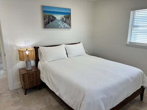 Une chambre à coucher comprenant un lit, une table de chevet, une lampe, un tableau représentant une scène de plage et une fenêtre avec des stores.