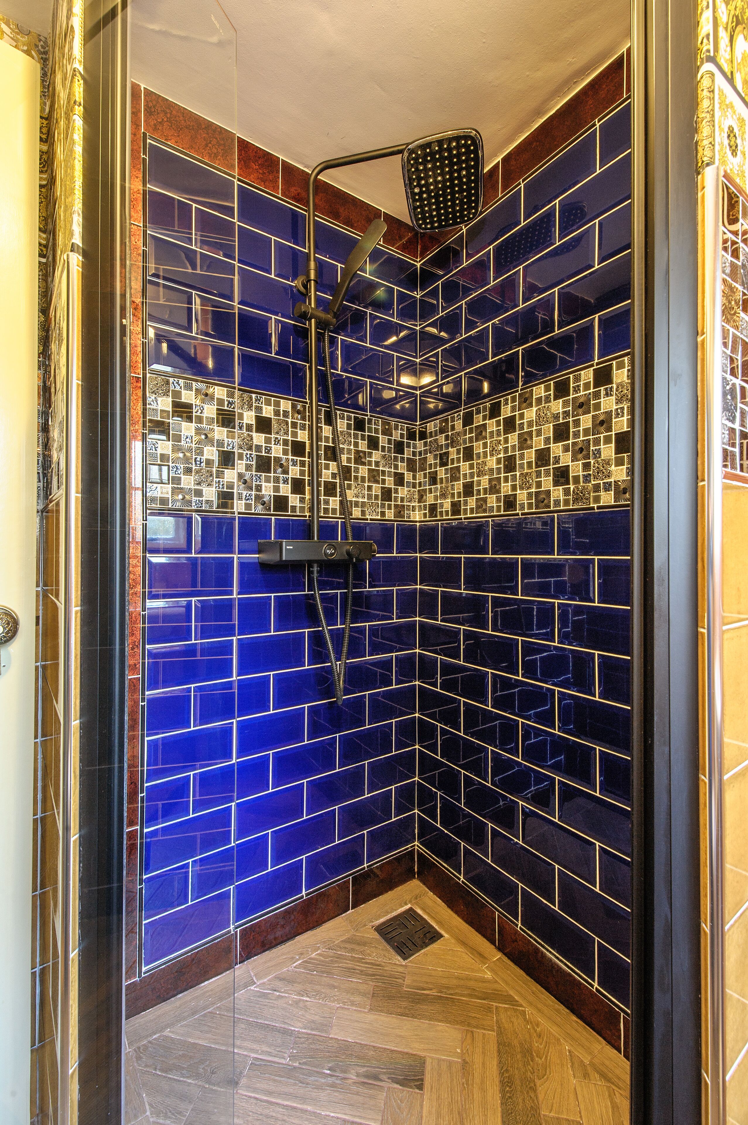 A shower with blue mosaic tiles and a wooden floor.