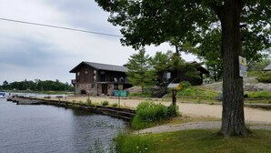Una cabaña junto a un lago con un muelle, una escalera que lleva a una terraza, y un cartel.