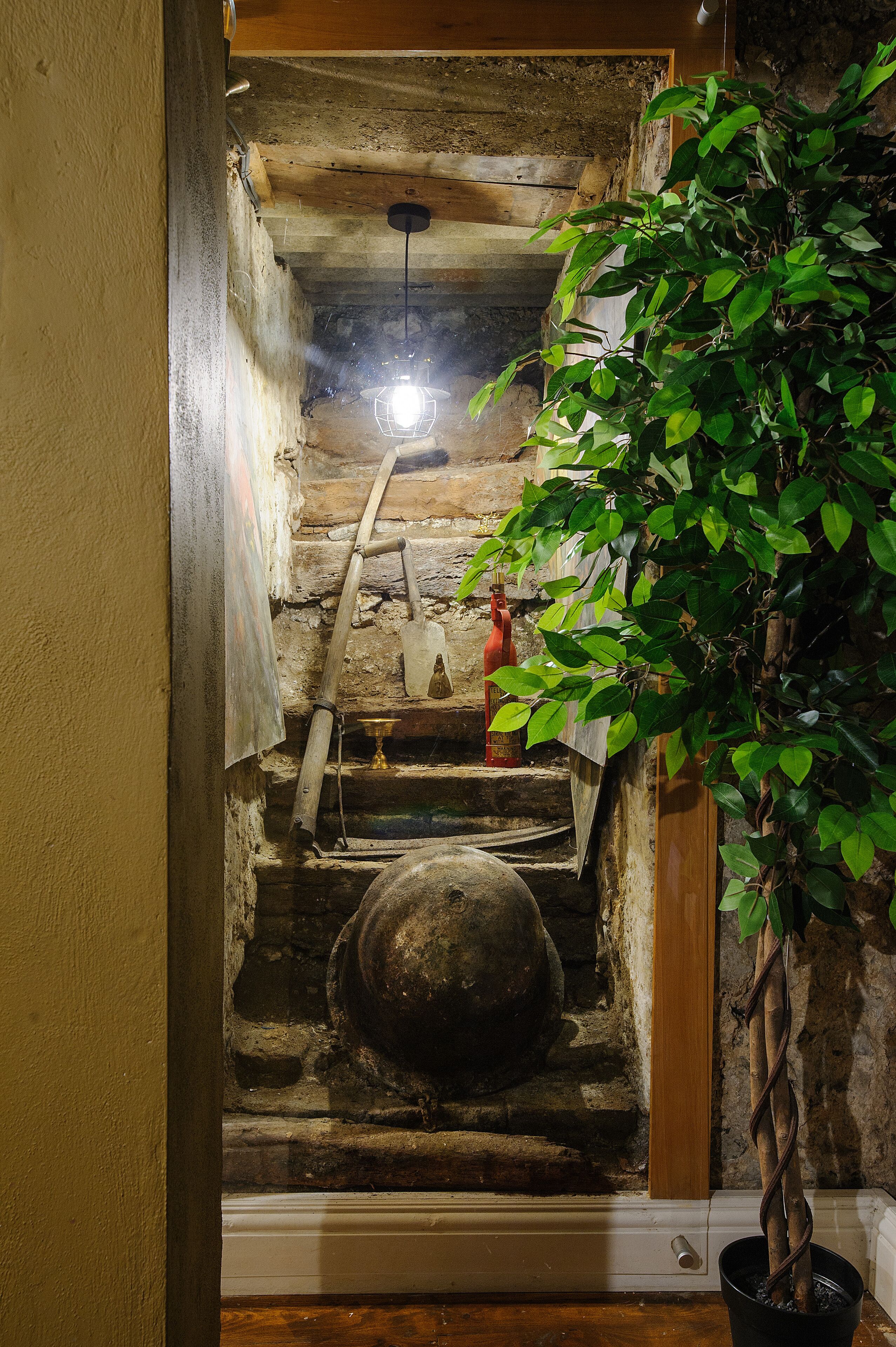 Uma escadaria estreita com corrimão de madeira, um vaso de planta à direita e uma parede de pedra.
