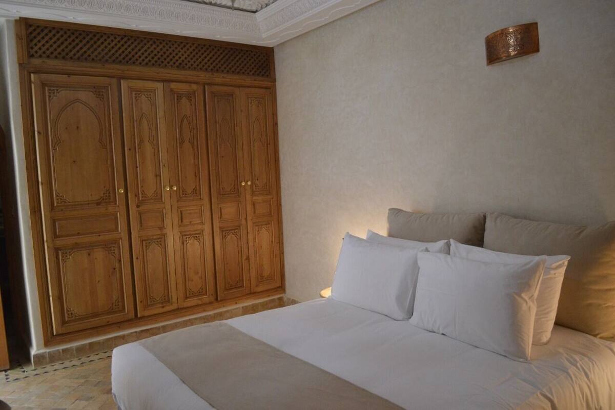 Un dormitorio con un amplio armario de madera, una cama con ropa de cama blanca y una mesita de noche pequeña.