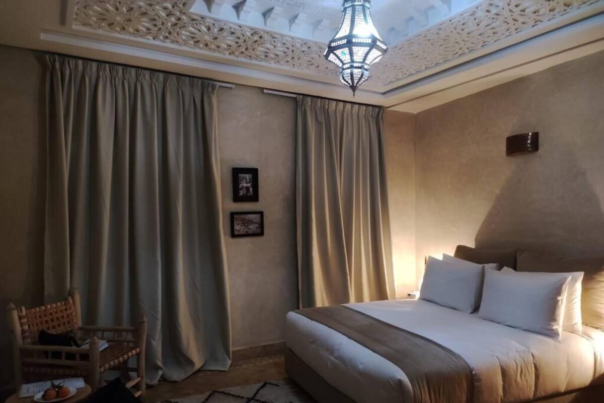 Habitación de hotel con una cama grande, una silla de madera, una mesita con una fuente de fruta y un techo decorativo.