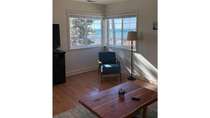 A living room with a wooden coffee table, a blue armchair, a TV, and large windows offering a view of the sea. - Fifth house from beach Avail June 14 - August18.  (Pismo Beach)