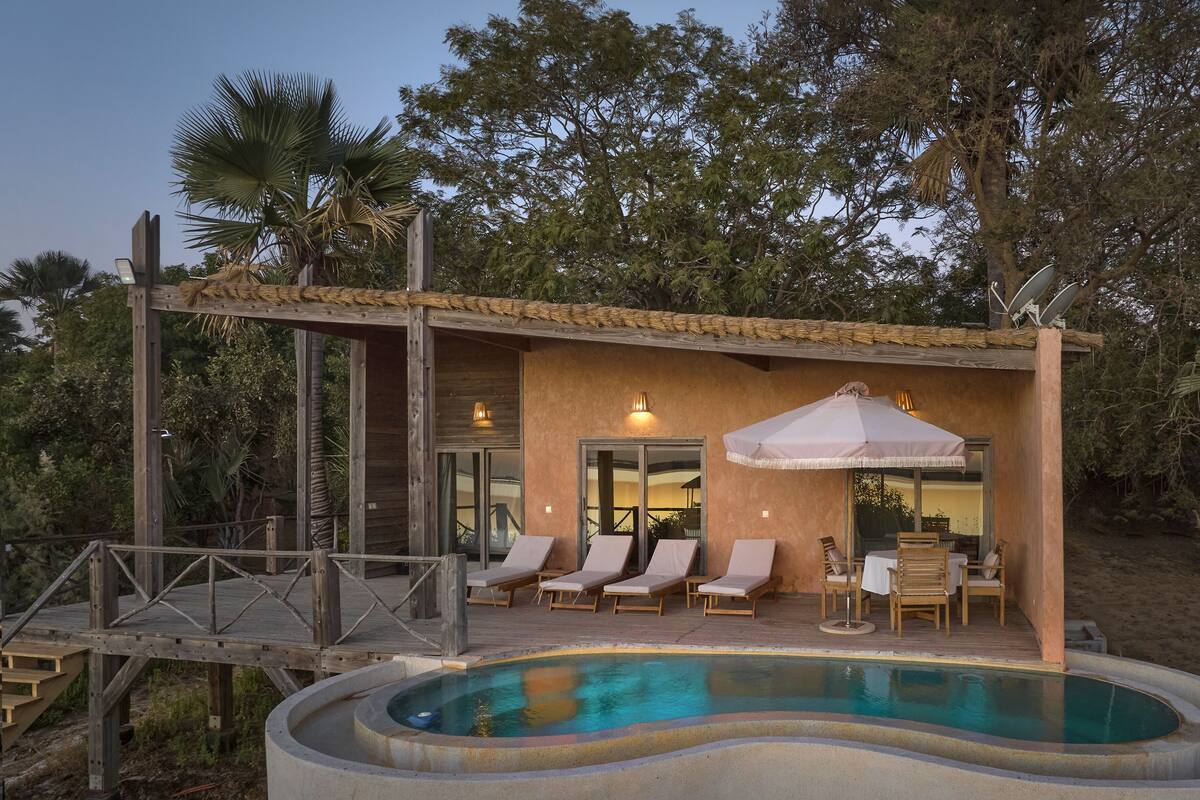 A poolside area with lounge chairs and a table, surrounded by a wooden deck and a covered seating area.