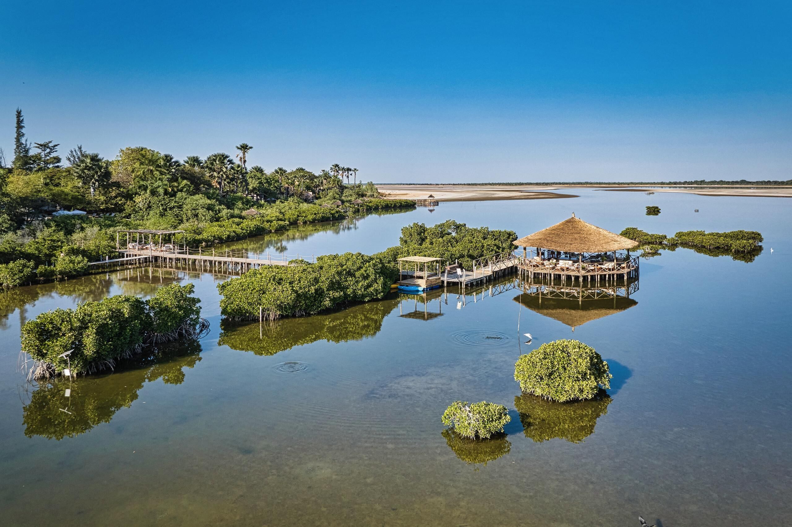 A tranquil water body with a wooden dock and a thatched-roof structure, surrounded by lush greenery.