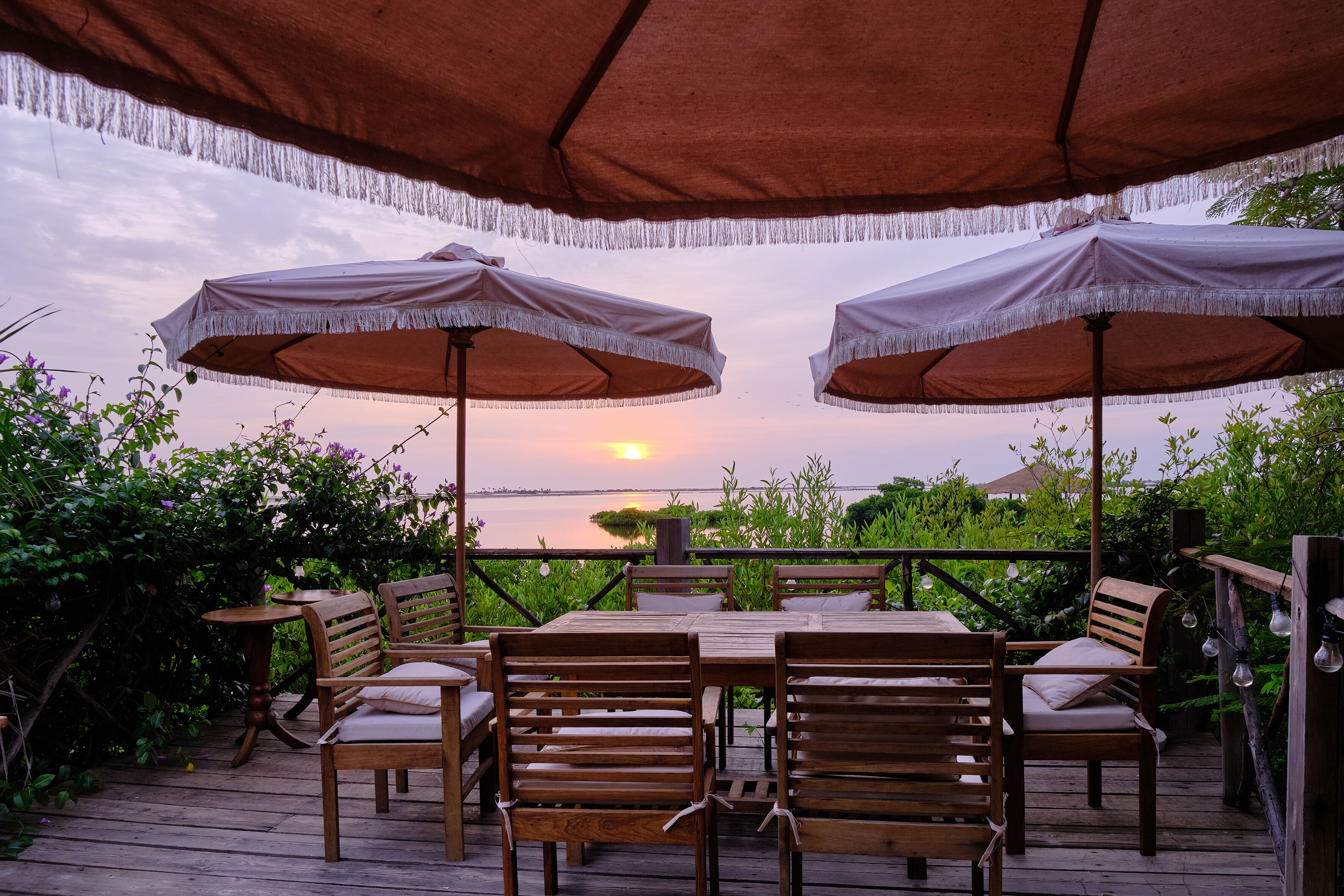An outdoor patio with wooden furniture and umbrellas, overlooking a body of water at sunset.