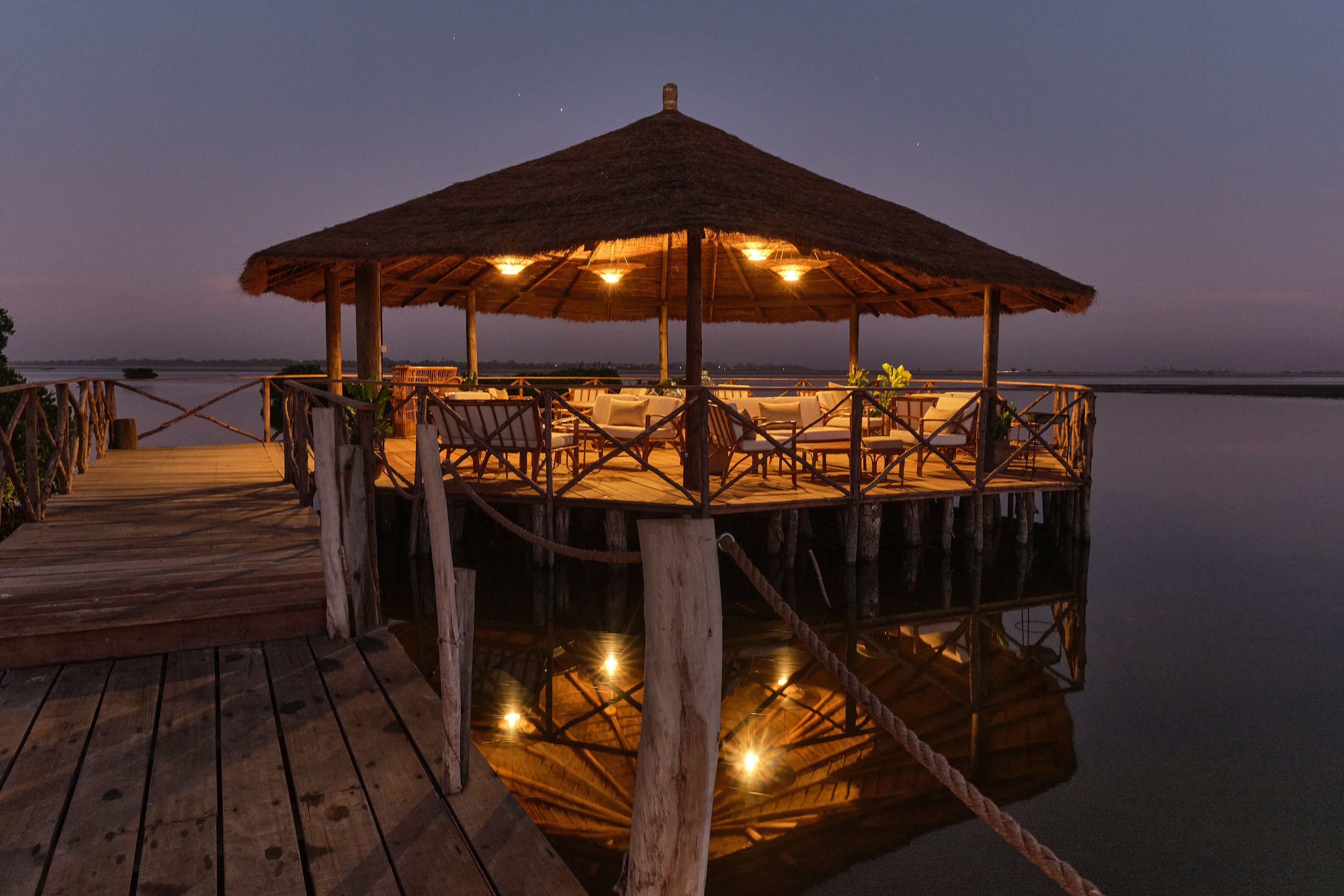 Una terraza de madera que conduce a una estructura con techo de paja y asientos, con vista a aguas tranquilas.