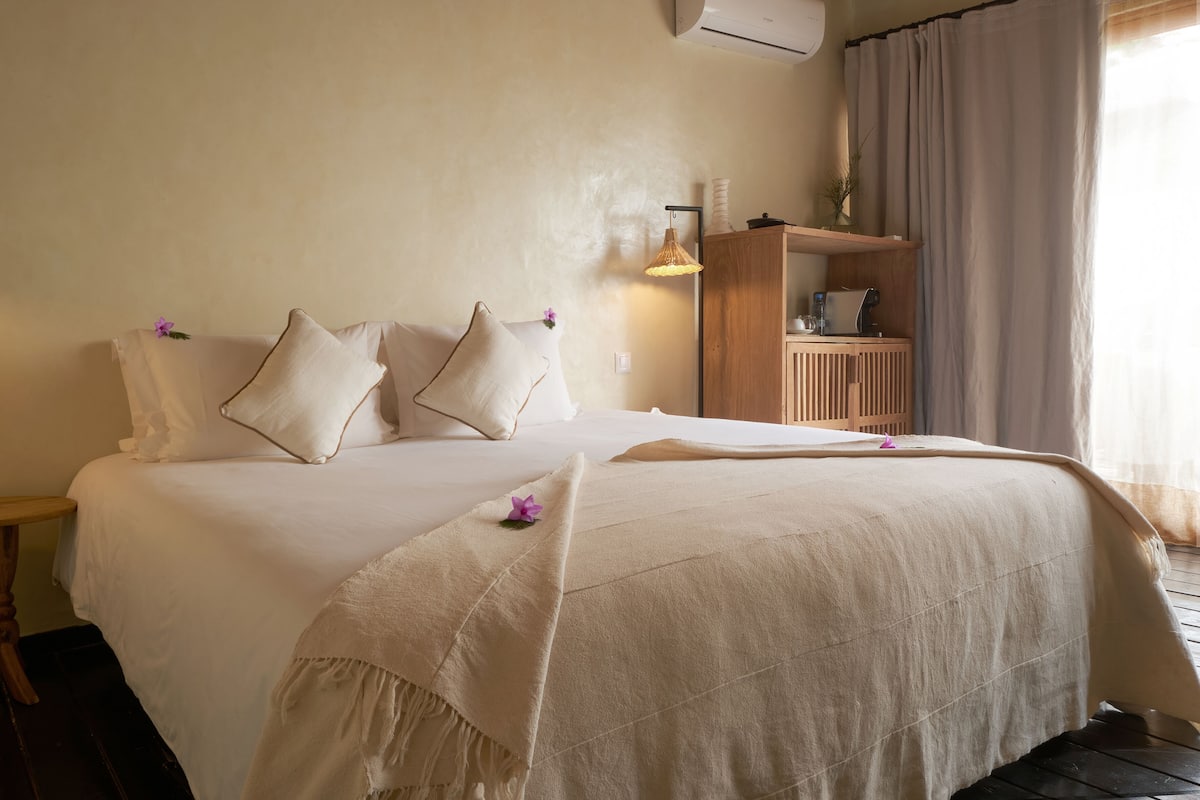 A neatly made bed with a beige bedspread and pillows, a bedside table with a lamp, and a wooden cabinet with a vase on top.