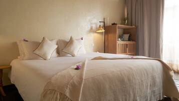 A neatly made bed with a beige bedspread and pillows, a bedside table with a lamp, and a wooden cabinet with a vase on top.