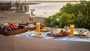 A breakfast setup with a view of a beach and a pier, including a wooden tray with watermelon, eggs, and pastries.