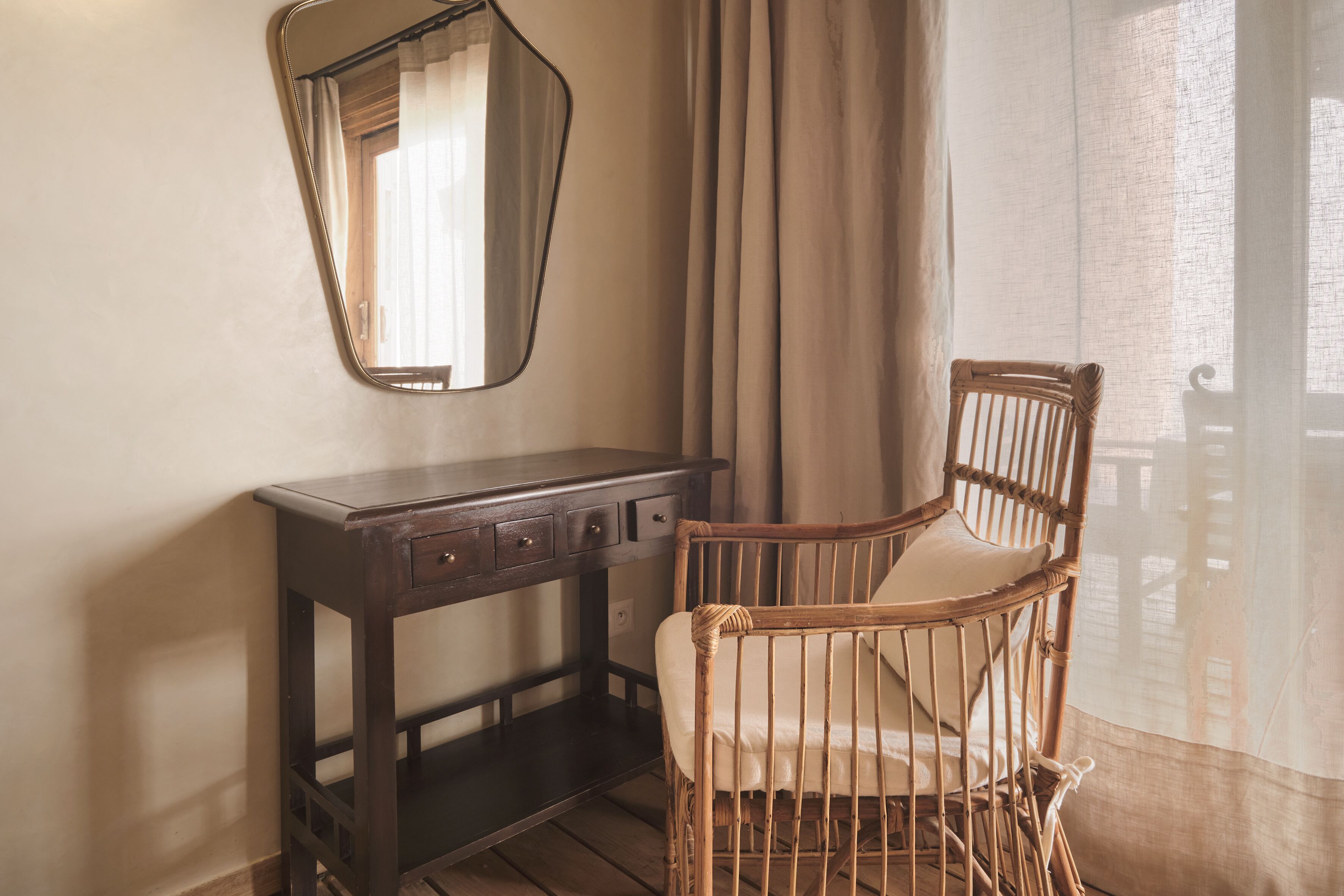 A room with a wooden desk, a wicker chair, a mirror, and a window with curtains.