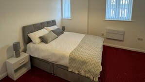 A bedroom with a bed, bedside tables, a heater, and two windows with blinds. - Apt Sleeps5-parkg-netflix-near City Centre/etihad (Manchester)