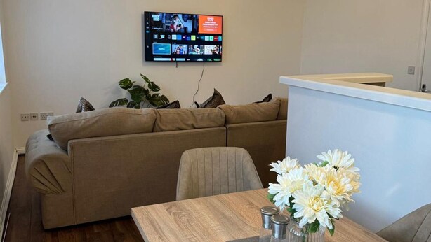A modern living room with a beige sofa, a wooden dining table with a vase of flowers, and a flat-screen TV mounted on the wall displaying various channels. - Apt Sleeps5-parkg-netflix-near City Centre/etihad (Manchester)
