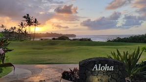 Um campo de golfe com uma placa de pedra que diz "Aloha" em primeiro plano, rodeado por vegetação exuberante e palmeiras.