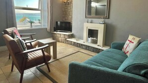 A living room with a stone fireplace, a television, a sofa, and a view of the ocean. - Min y Graig, New Quay (New Quay)