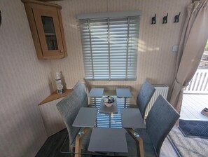 A compact living space with a glass-top table surrounded by chairs, a window with blinds, a small cabinet, and a radiator. - Beach House Dog Friendly - Anderby Creek (Skegness)