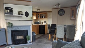 A modern living room with a fireplace, a dining area with chairs, and a kitchen in the background. - Beach House Dog Friendly - Anderby Creek (Skegness)