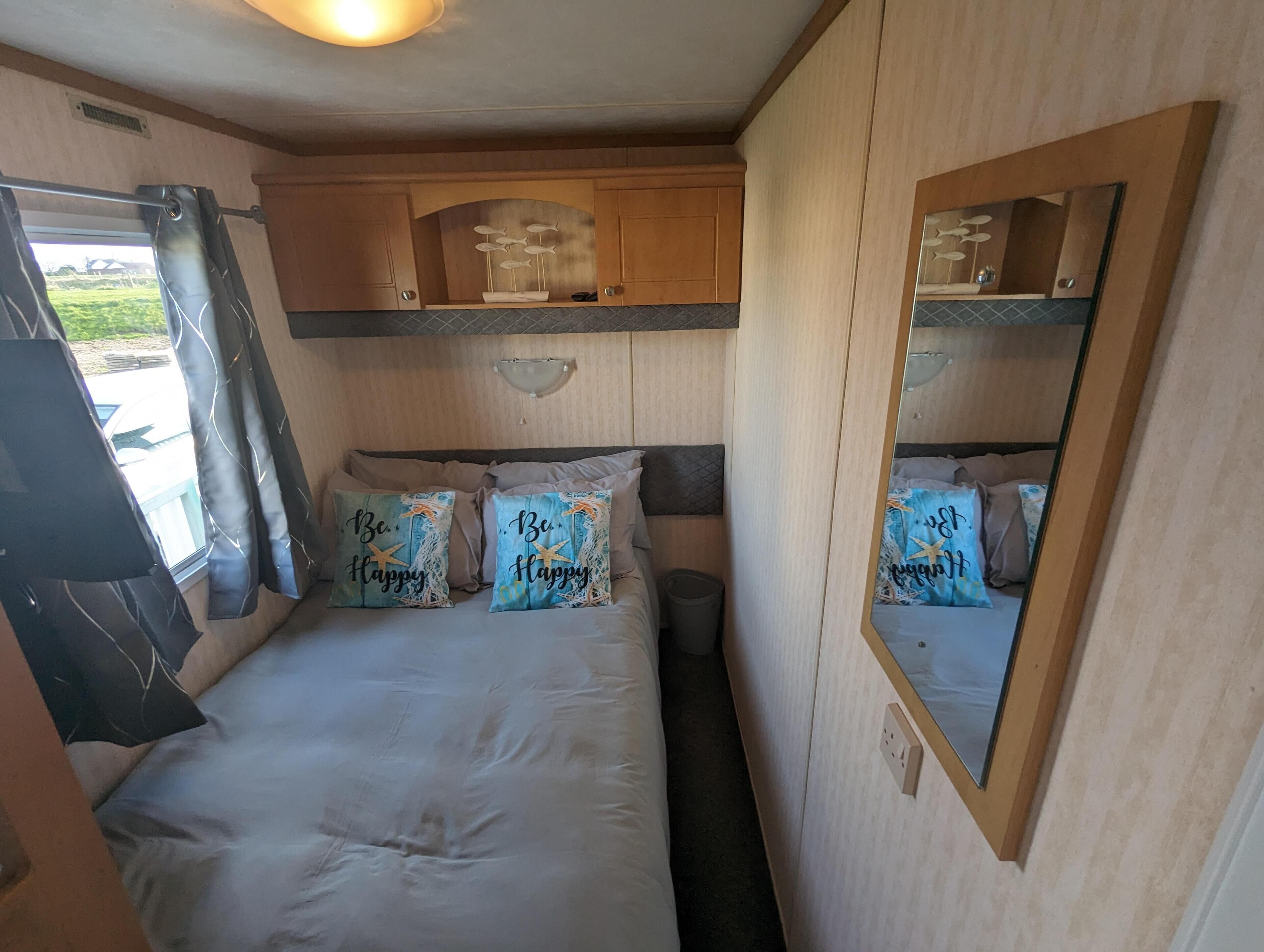 Een gezellige slaapkamer met een bed, twee kussens met de tekst 'Be Happy', een spiegel en houten kasten.