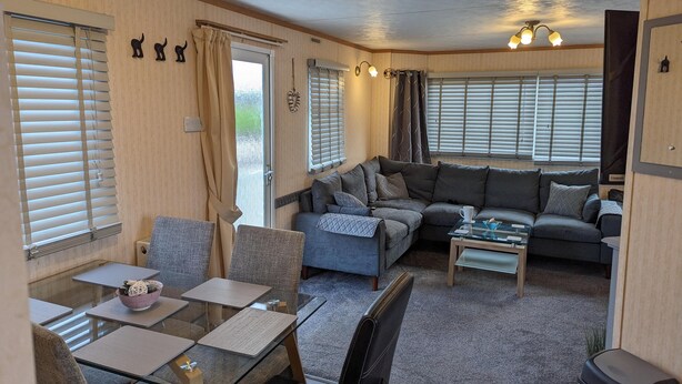 A cozy living room with a sofa, a coffee table, and a dining area with a glass table and chairs. - Beach House Dog Friendly - Anderby Creek (Skegness)