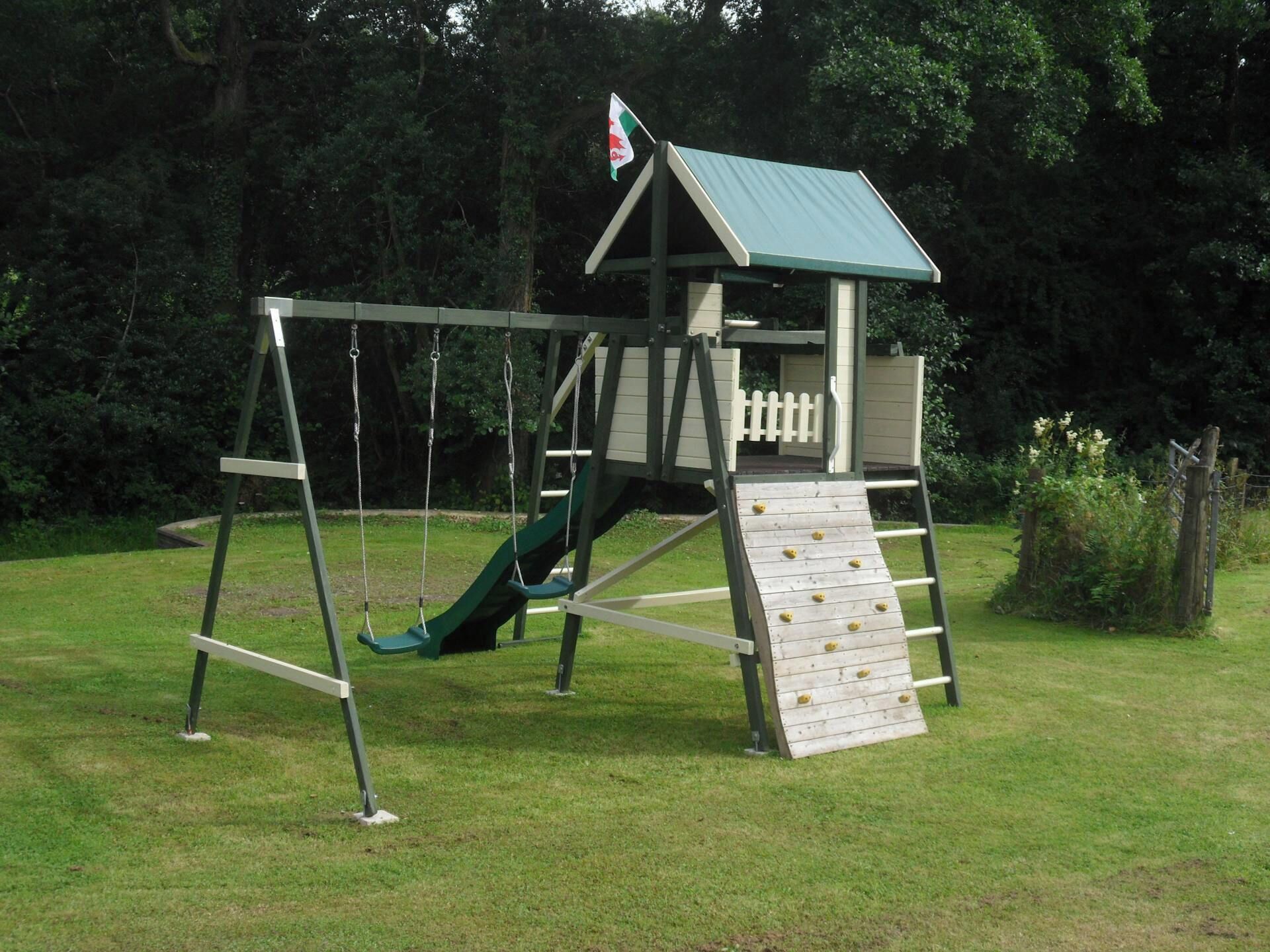 Een houten speelconstructie met een glijbaan en klimmuur, omringd door gras en bomen.