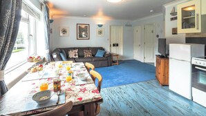 A cozy living room with a sofa, dining table with breakfast items, a kitchen with a refrigerator, and a blue carpet. - The Annexe, Llandysul (Penrhiw-llan)