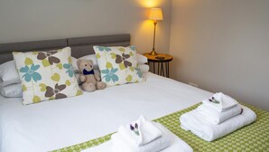 Una cama bien hecha con almohadones florales, un osito de peluche y toallas dobladas sobre un corredor de cama a cuadros verdes.