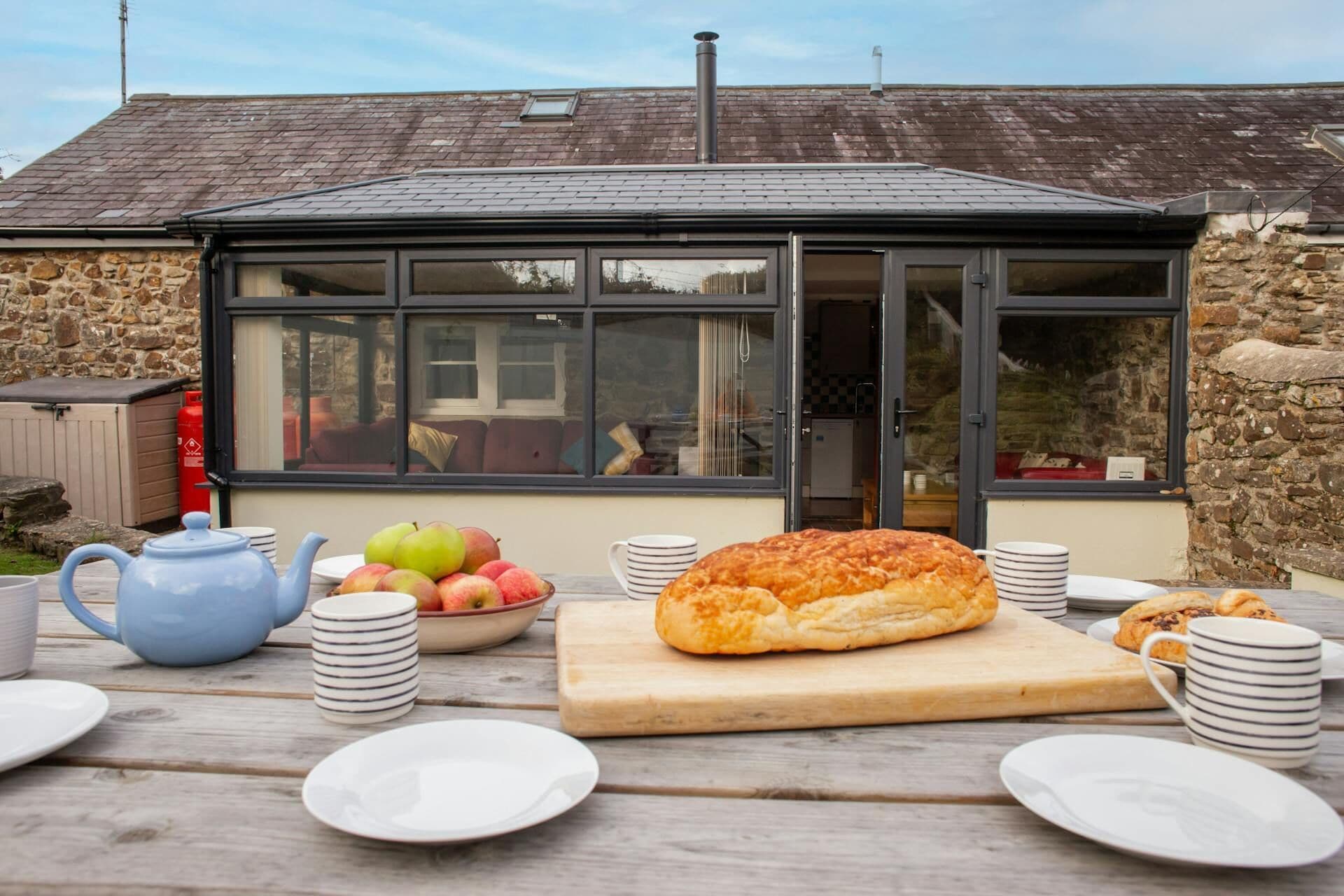 Une table en bois dressée pour le petit-déjeuner, avec une miche de pain, des fruits et une théière, placée devant un bâtiment en pierre doté d’une véranda en verre.