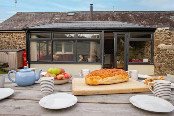 A breakfast setup with a loaf of bread, fruit, and tea on a wooden table in front of a stone building with a glass conservatory.