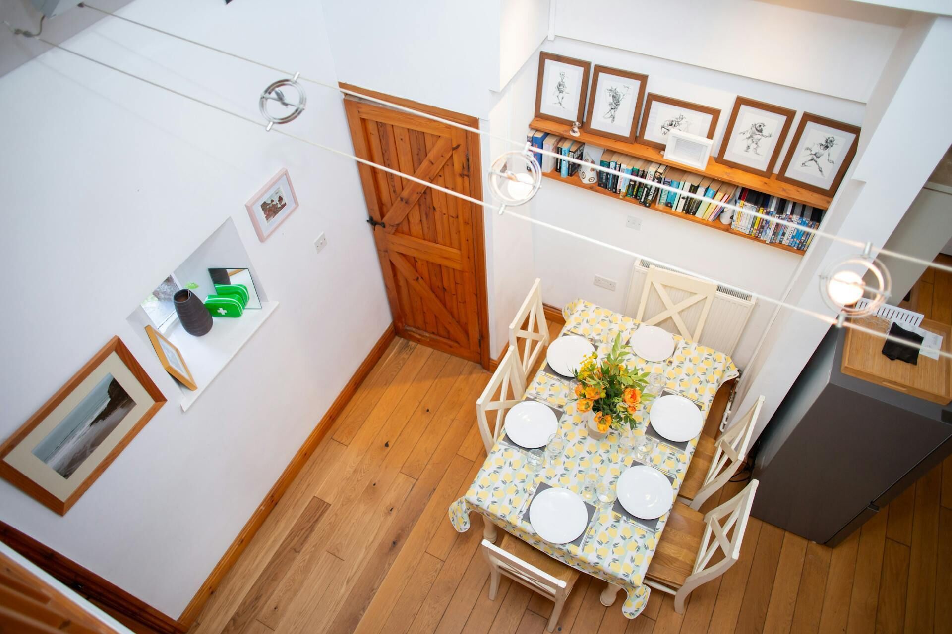 A compact kitchen with wooden flooring, a dining table set for four, and a bookshelf with framed artwork.