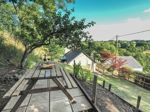 A picnic table with a bottle and glasses, surrounded by greenery and a house in the background. - Tanyfron, Llangrannog (Llwyndafydd)