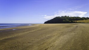 Uma extensa praia de areia com céu claro e uma colina com árvores e ruínas ao fundo.