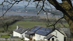 Uma casa com painéis solares no telhado, cercada por um quintal com cerca e vista para um rio e edifícios ao longe.