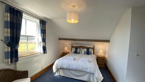 Um quarto com uma cama grande, duas mesas de cabeceira com abajures, uma cadeira de vime, uma janela com cortinas e um lustre.